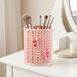 A pink plastic makeup brush holder with a knitted pattern and two small cartoon character decorations, holding several makeup brushes on a white vanity table.