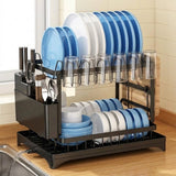A black metal kitchen dish rack set on a wooden countertop, holding blue, white, and gray plates, clear drinking glasses, and utensils. The rack is shown in a kitchen setting with a tiled wall and sink visible.