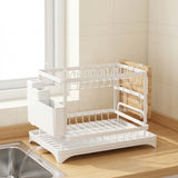 A white plastic dish drying rack with a sink basket and hanging hooks, shown on a wooden kitchen counter next to a sink in a tiled kitchen.