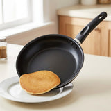 A black non-stick frying pan with a handle, shown tilted over a white plate holding a golden-brown pancake on a kitchen counter.