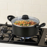 A black cooking pot with a clear glass lid and black handles sits on a lit gas stove burner in a kitchen setting, with pasta and vegetables visible inside.