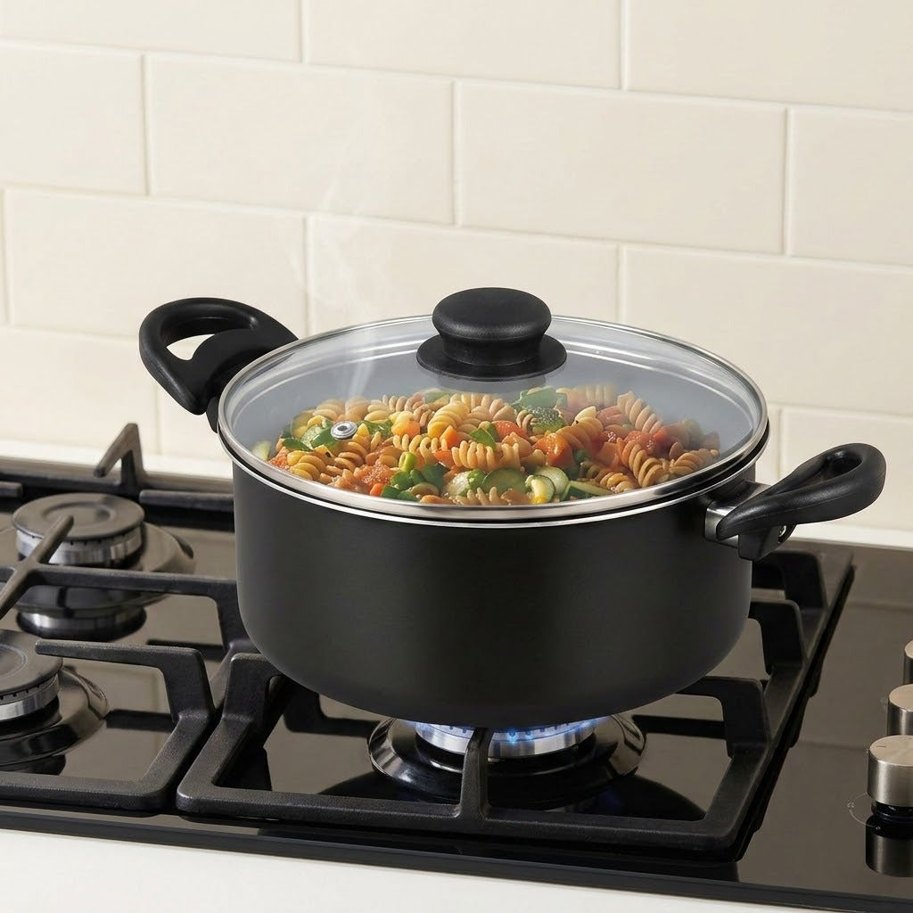 A black cooking pot with a clear glass lid and black handles sits on a lit gas stove burner in a kitchen setting, with pasta and vegetables visible inside.
