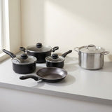 A 5-piece nonstick cookware set on a white countertop near a window. The set includes a stainless steel pot with a glass lid, two black saucepans with glass lids, and a black frying pan with a handle, all with black knobs.