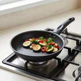 A black non-stick frying pan with a handle sits on a gas stovetop, cooking sliced zucchini, broccoli, and red bell peppers.