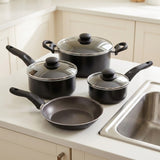 A 4-piece non-stick cookware set in black with clear glass lids and black handles, arranged on a white kitchen countertop next to a stainless steel sink.