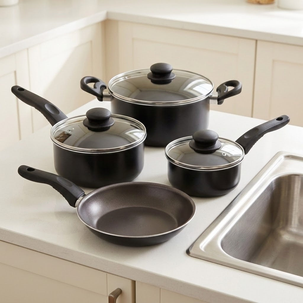 A 4-piece non-stick cookware set in black with clear glass lids and black handles, arranged on a white kitchen countertop next to a stainless steel sink.