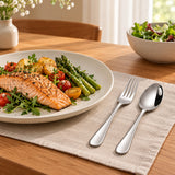 Matching stainless steel spoon and fork beside a plated lunch on a warm dining table.