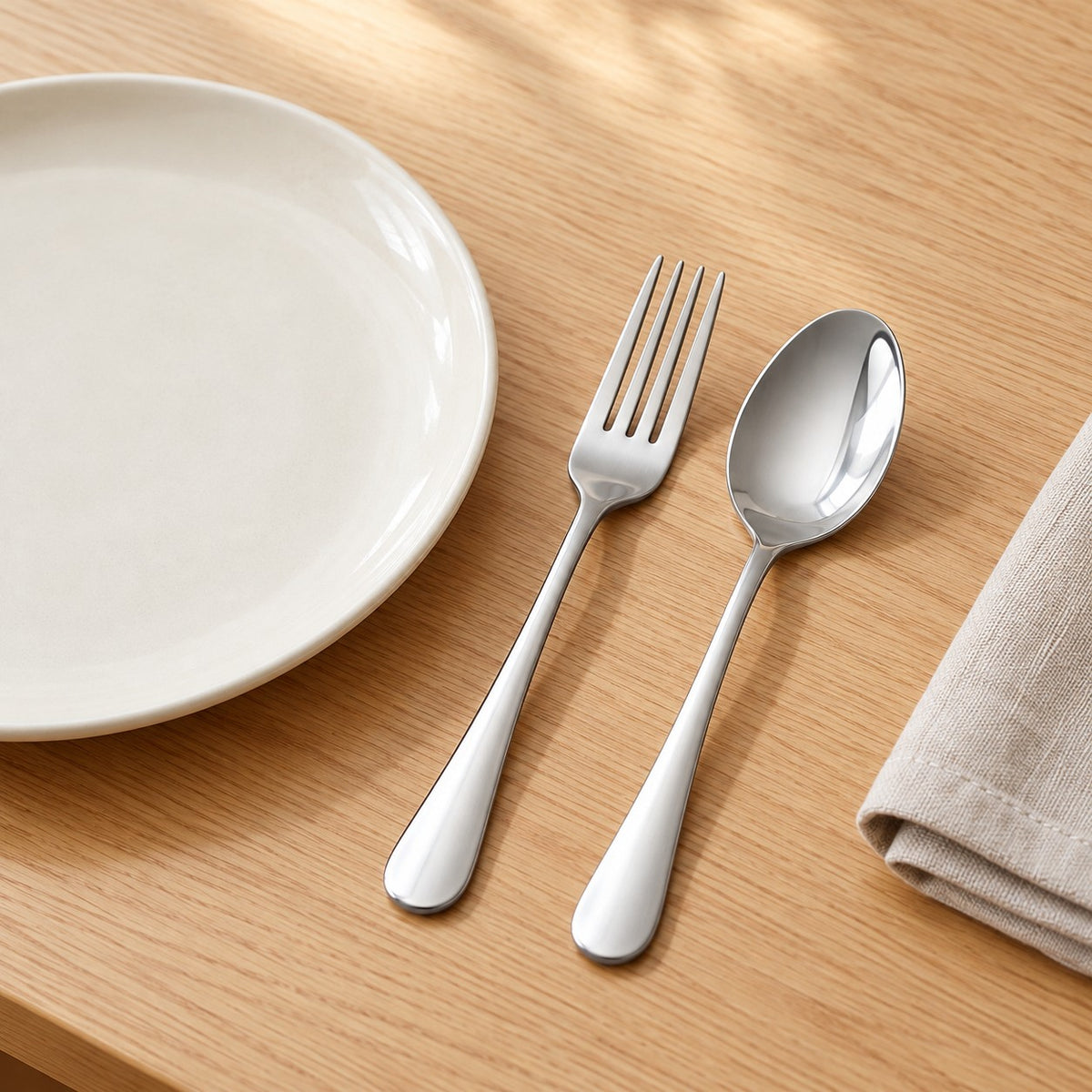 Stainless steel spoon and fork beside a dinner plate show their everyday table setting size.