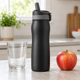 Black 24 oz insulated straw bottle stands beside a drinking glass and apple on a kitchen counter for scale.