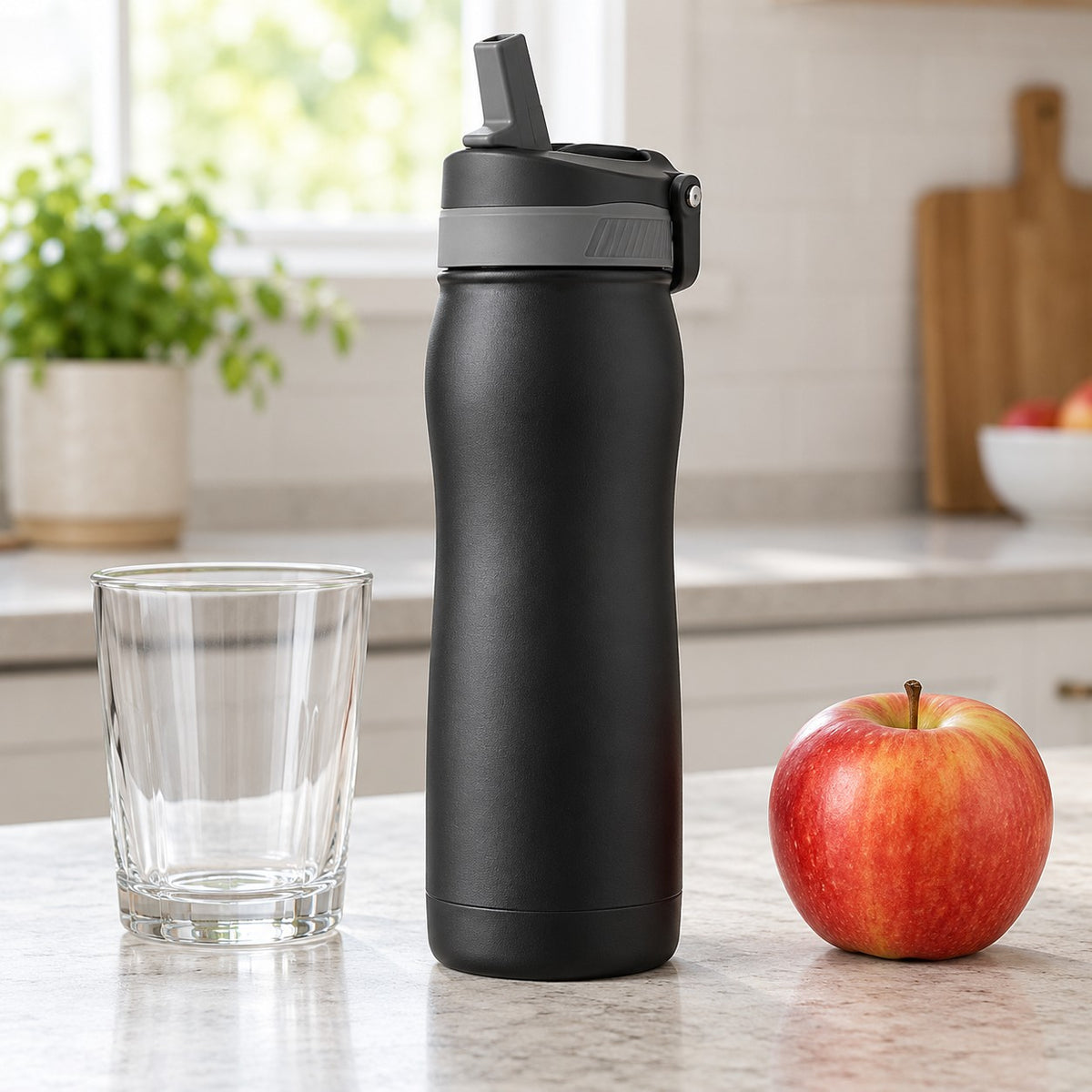Black 24 oz insulated straw bottle stands beside a drinking glass and apple on a kitchen counter for scale.