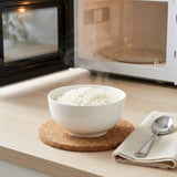 A white ceramic bowl filled with white rice sits on a round cork coaster on a light wood countertop, with a folded beige napkin and spoon beside it and a microwave in the background.