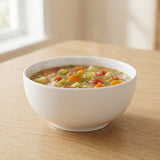 A white ceramic bowl filled with vegetable soup sits on a light wood table in a softly lit indoor setting.
