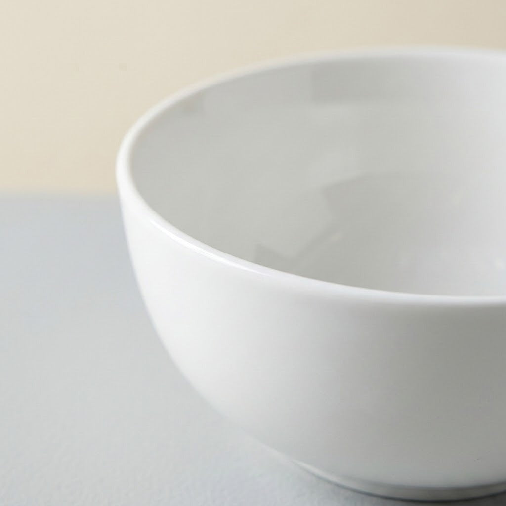 A close-up of a white ceramic bowl with a smooth, matte finish, set against a neutral light gray and beige background.