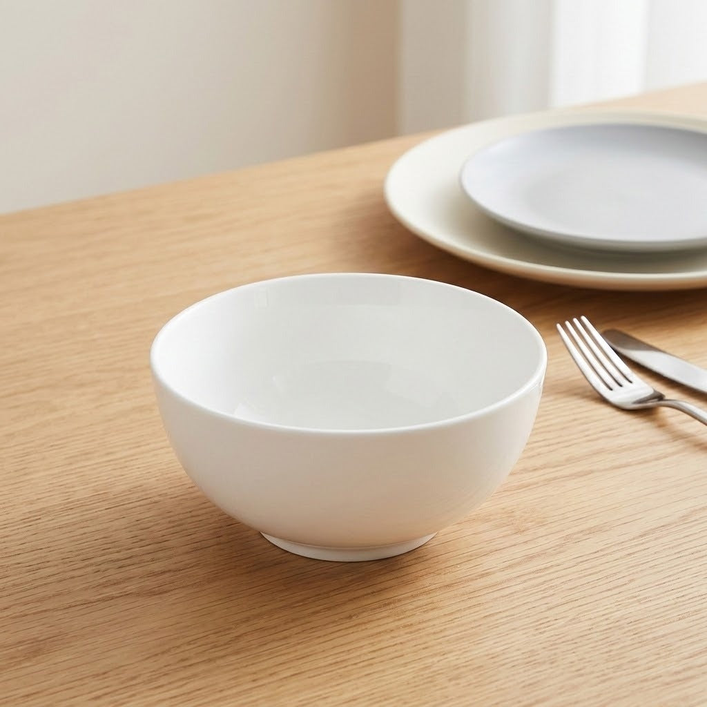 A white ceramic bowl sits on a light wood table, with matching plates and a fork blurred in the background.