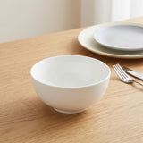 A white ceramic bowl sits on a light wood table, with matching plates and a fork blurred in the background.