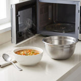 A stainless steel mixing bowl sits on a kitchen counter next to a white bowl of soup and a spoon, with an open microwave in the background.