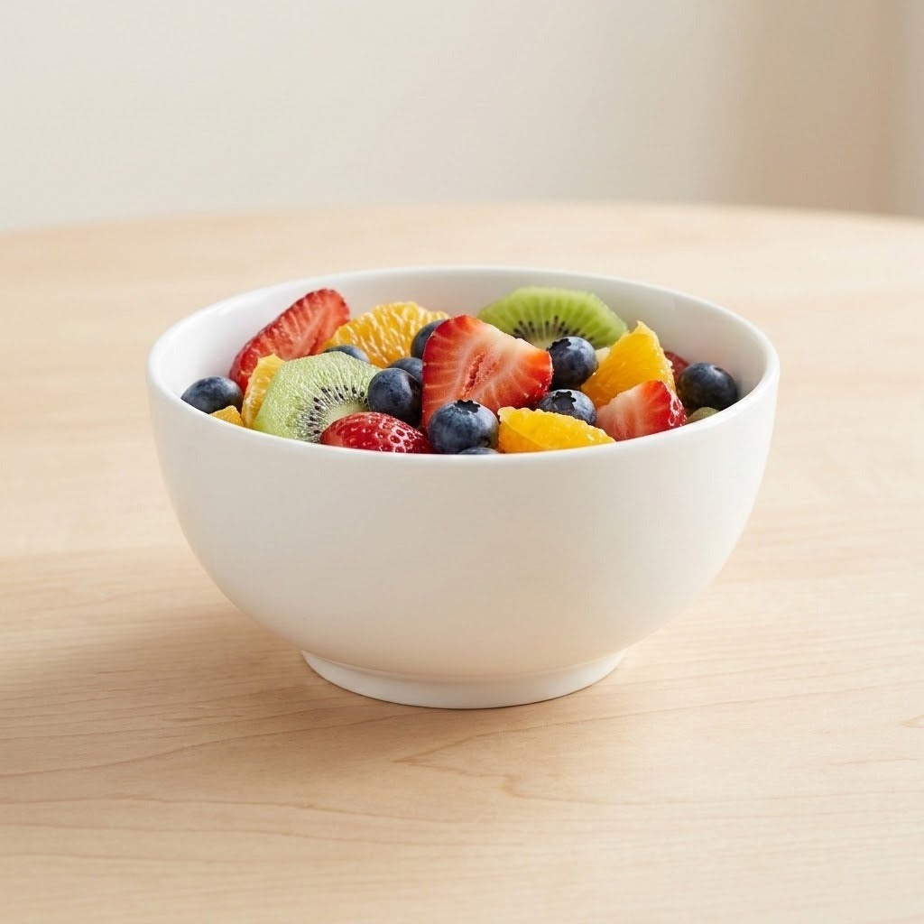 A white ceramic bowl filled with fresh fruit, including strawberries, blueberries, kiwi, and orange slices, set on a light wood table.