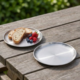 A round silver metal plate sits on a weathered wooden table outdoors, with another plate holding bread slices and berries placed nearby.