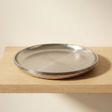 Three stacked round stainless steel plates with a brushed finish, resting on a light wood shelf against a beige wall.
