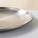A close-up of a brushed stainless steel round tray or plate, showing its metallic texture and curved rim against a neutral light background.