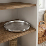 Two round stainless steel pizza pans stacked on a rustic wooden shelf in a kitchen setting.