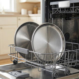Two large stainless steel round pans resting in the silver wire dishwasher basket inside an open dishwasher, with a blurred kitchen interior in the background.