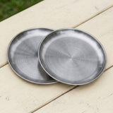 Two round brushed metal trays or shallow plates overlapping on a light wood surface outdoors.