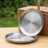 Two round brushed stainless steel serving platters, one standing upright and one lying flat, displayed on a wooden table with a wicker basket and blurred green garden background.