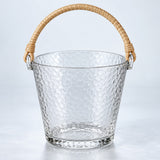 Clear Glass ice bucket with hammered texture and woven rattan handle on a pale backdrop.