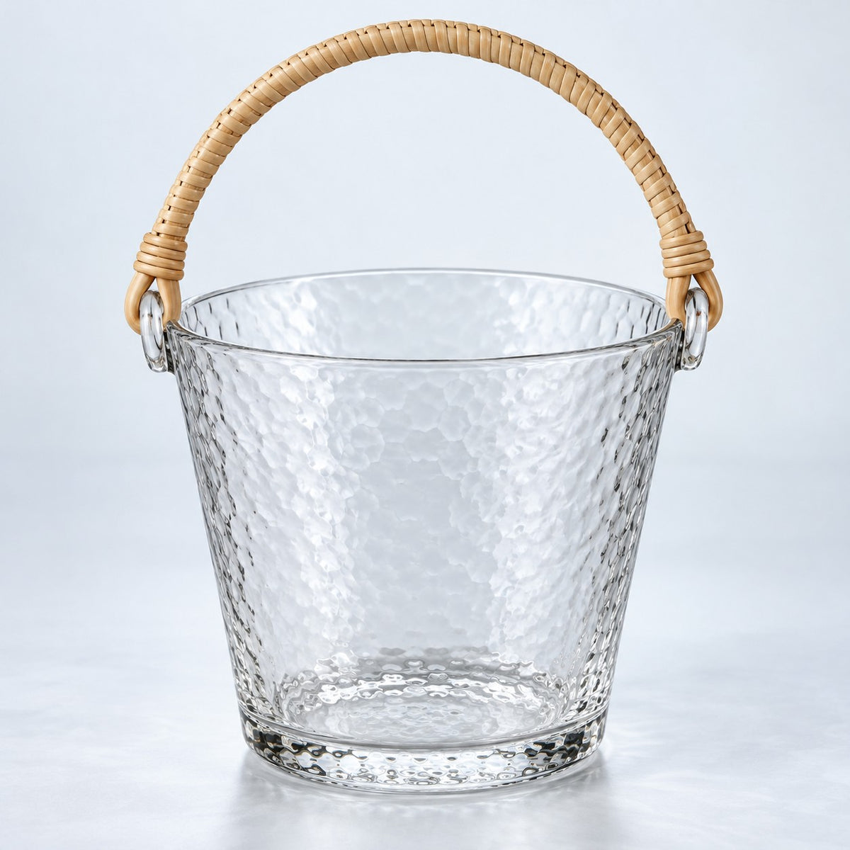 Clear Glass ice bucket with hammered texture and woven rattan handle on a pale backdrop.