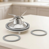 A stainless steel blender blade assembly with a gray rubber seal and two matching gray rings, displayed on a light beige countertop with a blurred kitchen background.