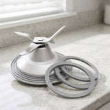 A stainless steel blender blade assembly with a four-blade impeller resting on a circular base, shown on a speckled kitchen countertop with two gray rubber seals in front.