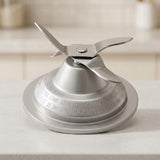 A stainless steel blender blade assembly with a four-blade impeller mounted on a circular metal base, shown on a light countertop with a softly blurred kitchen background.