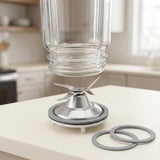 A clear glass jar with a metal blade assembly and gray rubber rings, shown on a light countertop in a softly blurred kitchen setting.