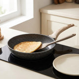 A dark speckled non-stick frying pan with a light wood handle sits on a black induction cooktop in a bright kitchen. A pancake is cooking in the pan, and a matching light wood spatula rests inside the pan.