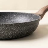 A close-up of a dark gray non-stick frying pan with white speckles and a light brown wooden handle, set against a warm off-white background.