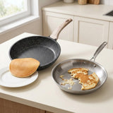 Two frying pans on a kitchen counter, one dark gray speckled pan with a wooden handle holding a pancake on a plate, and one stainless steel pan with a broken pancake inside.