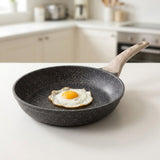 A dark gray speckled non-stick frying pan with a light wood handle sits on a white kitchen counter, containing a fried egg.