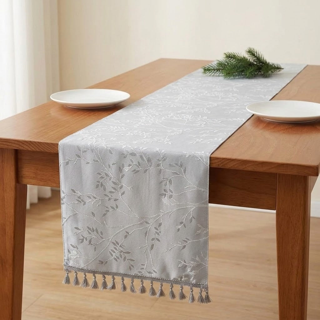 A light gray table runner with a subtle floral pattern and tassel fringe, styled on a wooden dining table with white plates and a green pine sprig.