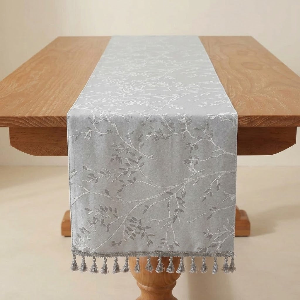 A light gray table runner with a white embroidered leaf and branch pattern, draped over a wooden dining table with matching tassels at the end.