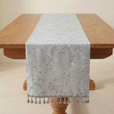 A light gray table runner with a white embroidered leaf and branch pattern, draped over a wooden dining table with matching tassels at the end.