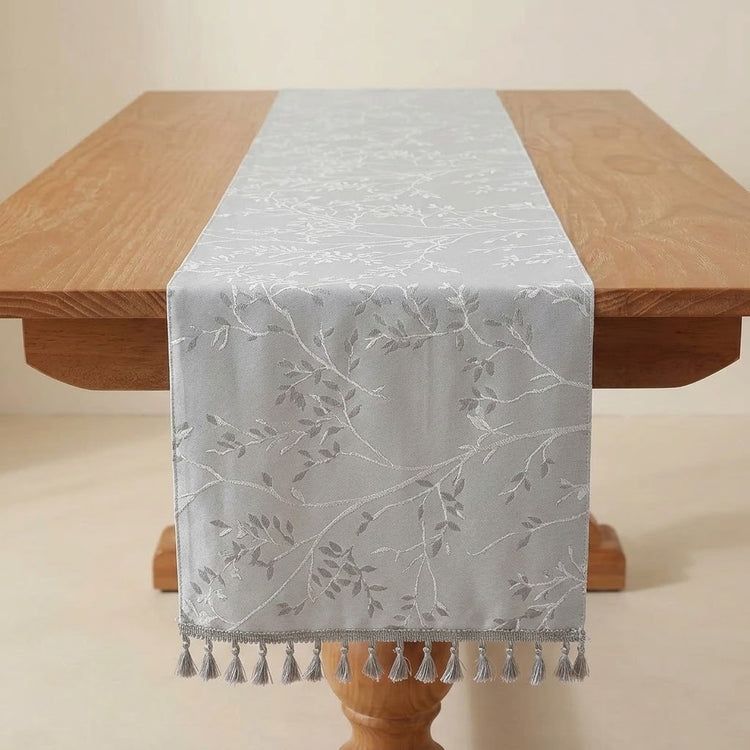 A light gray table runner with a white embroidered leaf and branch pattern, draped over a wooden dining table with matching tassels at the end.