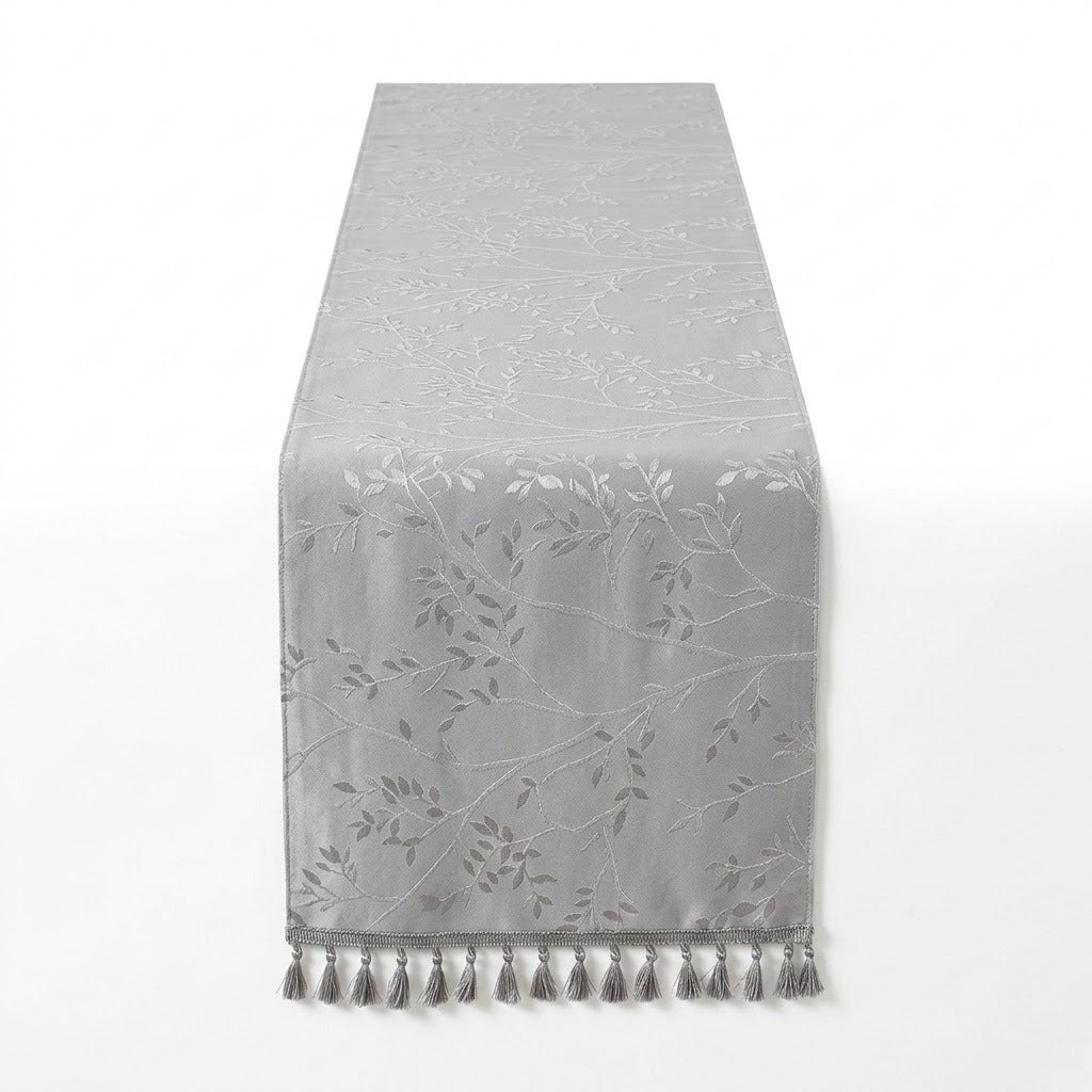 A light gray table runner with a subtle tonal floral and leaf pattern and matching tassel fringe along the bottom edge, shown against a plain white background.