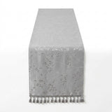 A light gray table runner with a subtle tonal floral and leaf pattern and matching tassel fringe along the bottom edge, shown against a plain white background.