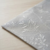 A close-up of a light gray fabric napkin or table linen with white embroidered floral and leaf patterns, resting on a light wood surface.