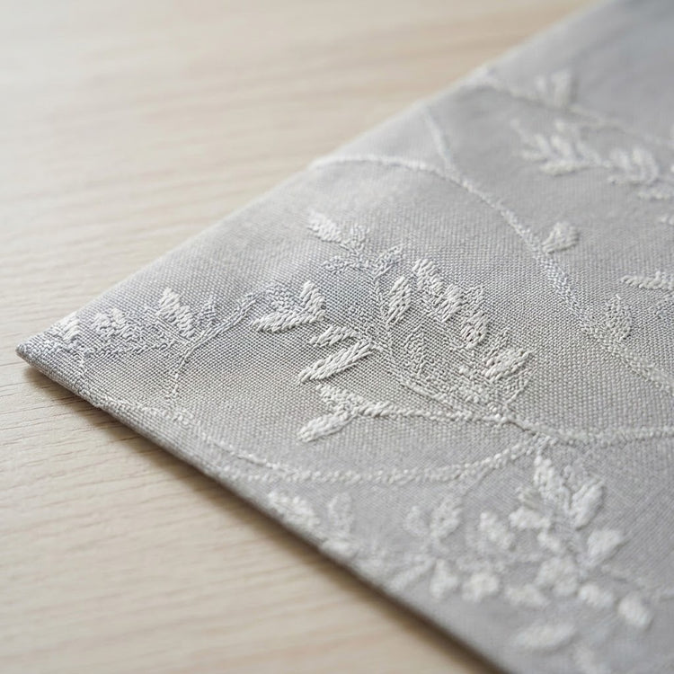 A close-up of a light gray fabric napkin or table linen with white embroidered floral and leaf patterns, resting on a light wood surface.