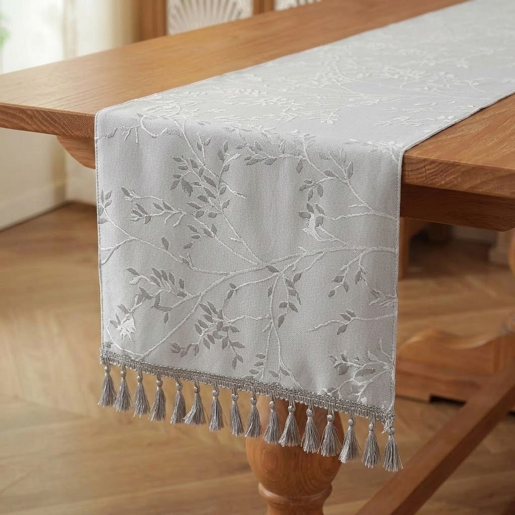 A light gray table runner with embroidered leafy branches and a tassel fringe, draped over a wooden dining table.