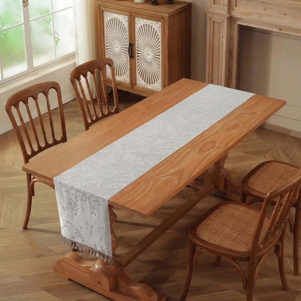 A light gray floral lace table runner with fringe is draped over a wooden dining table, surrounded by four wooden chairs in a warm, rustic dining room setting.