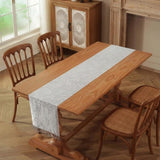 A light gray floral lace table runner with fringe is draped over a wooden dining table, surrounded by four wooden chairs in a warm, rustic dining room setting.