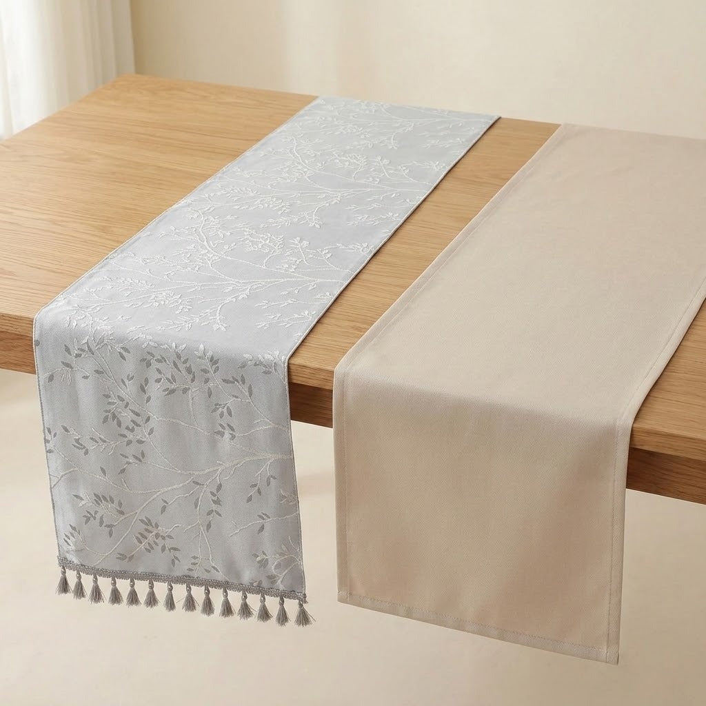 Two table runners displayed on a light wood table, one in pale blue-gray with a subtle floral vine pattern and tassel fringe, and one in solid cream.
